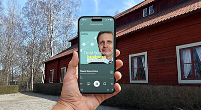En hand håller en iPhone som visar Spotify-podden "Business och barriärer" av David Stiernholm, med en traditionell röd svensk byggnad och kala björkträd i bakgrunden.