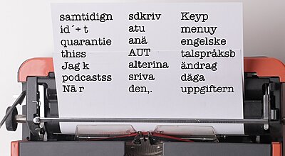 En skrivmaskin håller ett pappersark med fragmenterade svenska ord och fraser arrangerade i tre kolumner.
