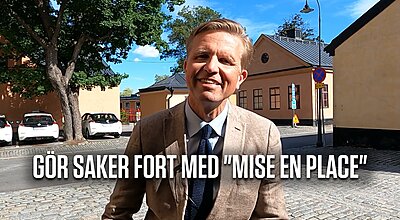 En leende man i en beige kostym och marinblå slips står på en kullerstensgata med den svenska texten "GÖR SAKER FORT MED 'MISE EN PLACE'" visad längst ned i bilden.
