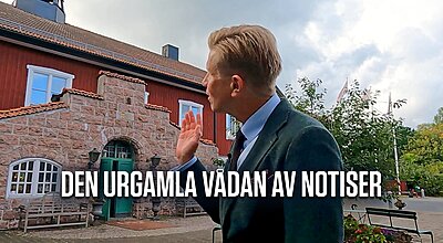 En man i mörk kostym gestikulerar med handen utanför en traditionell röd och stenbyggnad i skandinavisk stil, med den svenska texten "Den urgamla vådan av notiser" synlig i bilden.