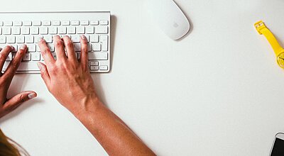 One and a half hand typing och a white keyboard on a white desk, where also there are a yellow wristwatch, a white mouse and a cellphone.