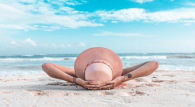 Händerna, armarna och hatten på en kvinna som ligger och njuter på en strand. I bakgrunden slår de turkosblå vågorna in.
