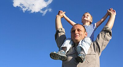 Ett litet barn sitter på en glad mans axlar och sträcker upp armarna mot en blå himmel