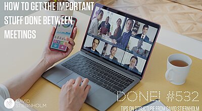 A laptop sits on a desk, showing a typical digital meeting. To the right is a white cup of tea. The meeting attendant's right hand rests on the keyboard while the left hand holds a smartphone.