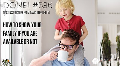 Brown haired man in black rimmed glasses drinking coffee wearing a light blue t-shirt while his young son sits on his shoulders.