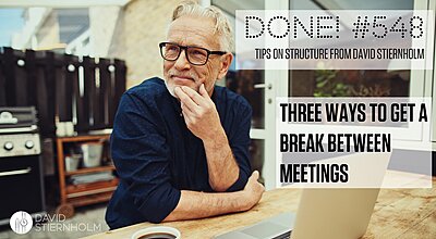 A white haired man in a short white beard and black rimmed glasses sits at a wooden desk, leaning his chin in his left hand. Dressed in an indigo shirt, he looks thoughtful. Before him, on the desk, sits a laptop. He is obviously taking a break from work for a few minutes.